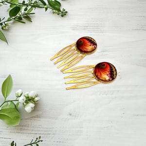 May include: Two gold-toned hair combs featuring circular cabochons with a vibrant fractal pattern in shades of red, orange, yellow, and deep purple. Each comb has four prongs and a smooth, polished metal finish, showcasing a swirling design.