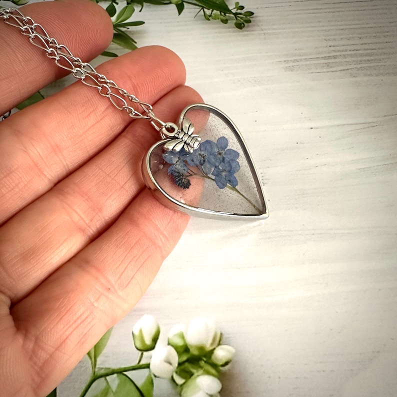 May include: A silver heart-shaped pendant necklace with a delicate chain. The pendant contains a pressed forget-me-not flower with a small silver bee charm.