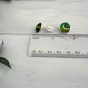 May include: A pair of silver-toned clip-on earrings featuring circular cabochons with a green and gold spiral shell pattern. The earrings are positioned next to a clear plastic ruler showing a diameter of approximately 0.5 inches for each piece.