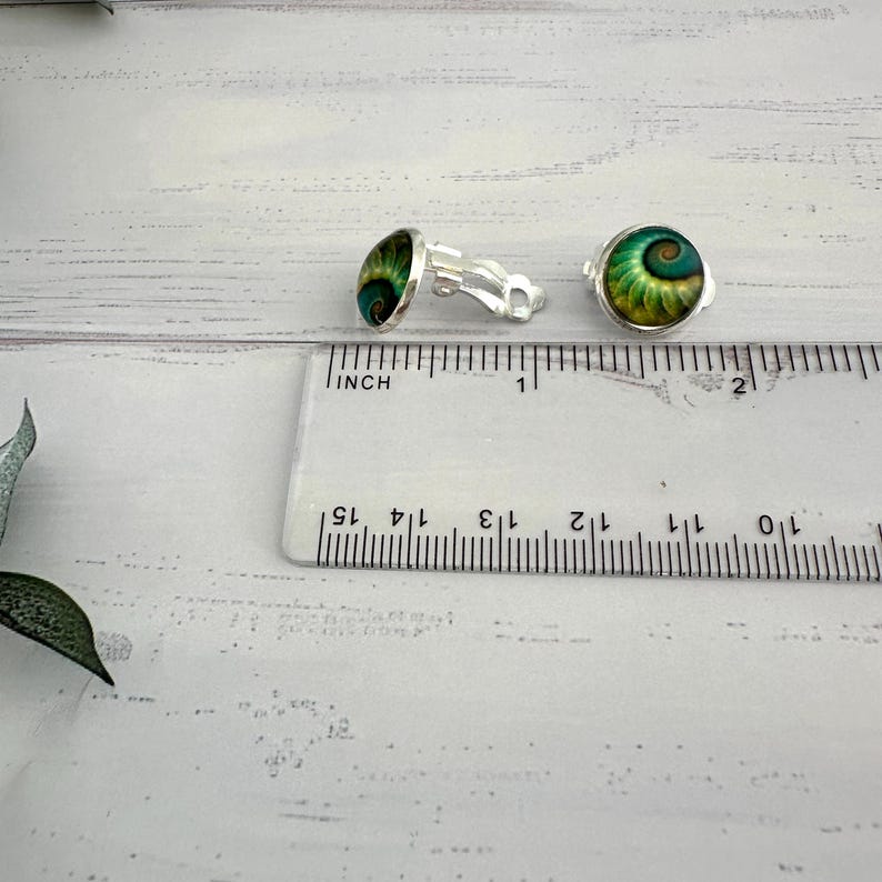 May include: A pair of silver-toned clip-on earrings featuring circular cabochons with a green and gold spiral shell pattern. The earrings are positioned next to a clear plastic ruler showing a diameter of approximately 0.5 inches for each piece.