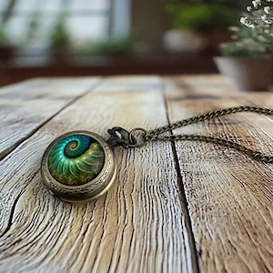 May include: Antique bronze pocket watch pendant with a spiral design in shades of green and blue. The watch hangs from a matching chain, creating a vintage aesthetic. The watch is displayed on a wooden surface.