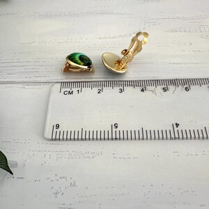 May include: A pair of gold-tone clip-on earrings featuring a round, green, and black swirl pattern. The earrings are positioned next to a clear plastic ruler, showing a diameter of approximately 0.5 inches for each earring piece.