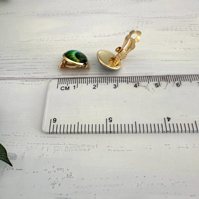 May include: A pair of gold-tone clip-on earrings featuring a round, green, and black swirl pattern. The earrings are positioned next to a clear plastic ruler, showing a diameter of approximately 0.5 inches for each earring piece.