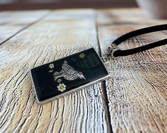 May include: Rectangular pendant with a black background, featuring a detailed silver bird design and small white daisies. The pendant is attached to a black cord necklace with a silver clasp. The pendant is approximately 1.5 inches long.
