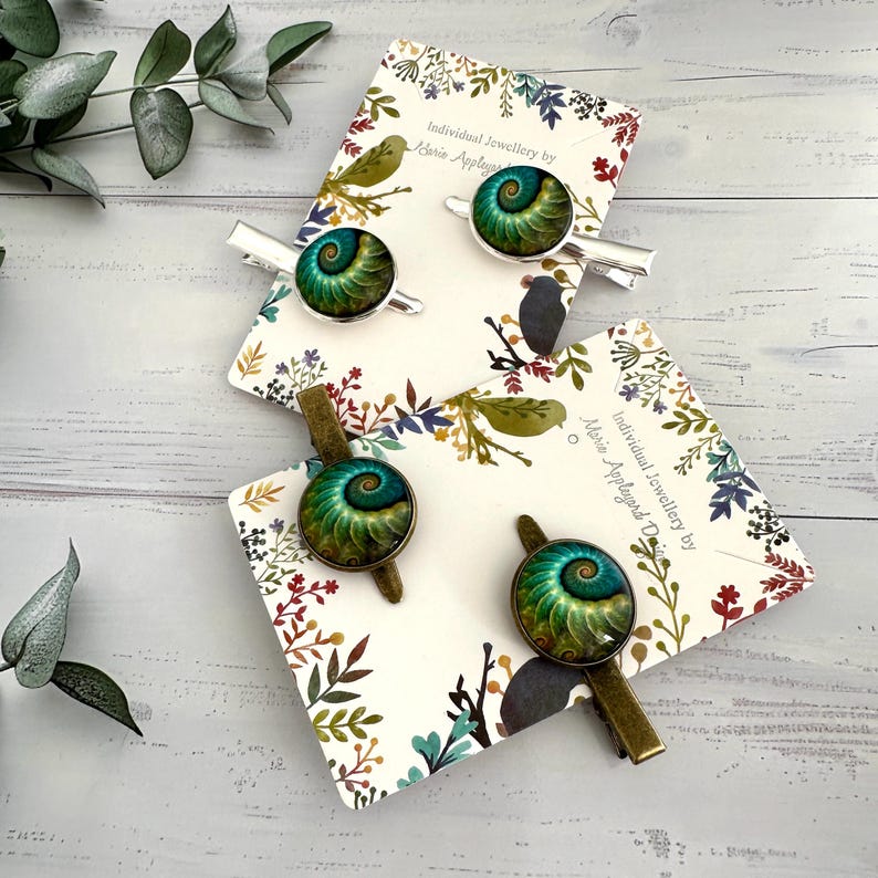 May include: Four hair clips with a green and blue spiral design, resembling a nautilus shell. The clips are on cards with floral patterns and the text "Individual Jewellery by Marie Applegard Design". The clips have silver and bronze finishes.