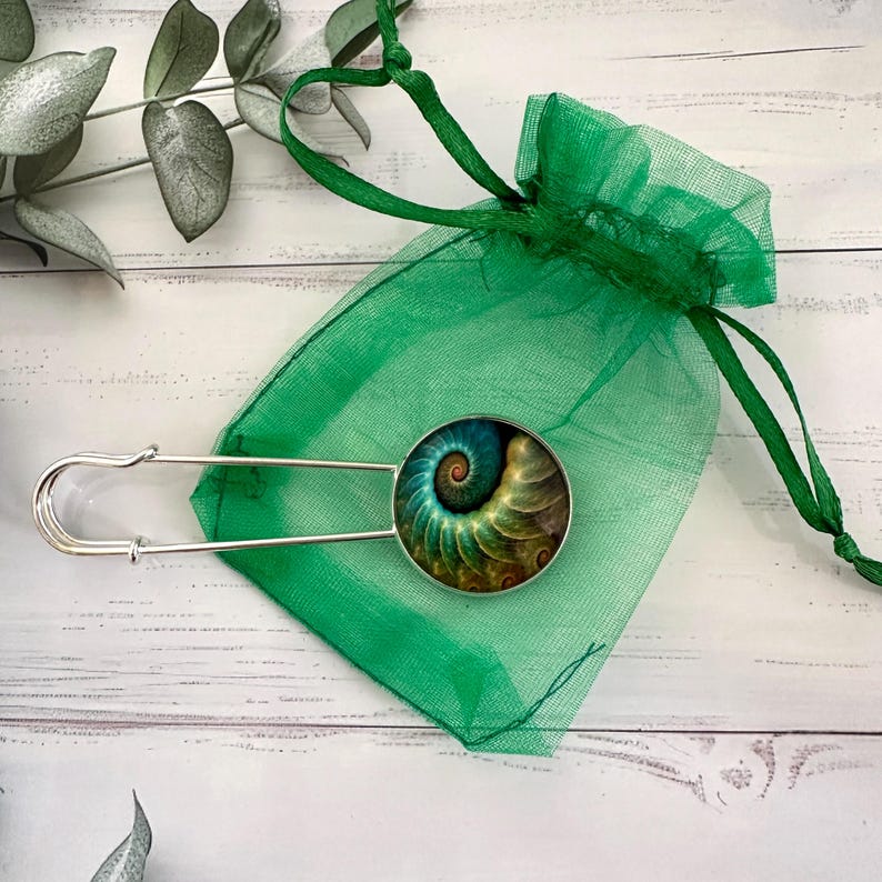 May include: A silver safety pin with a round, green, gold, and brown patterned glass cabochon. The pin is in a green mesh bag.