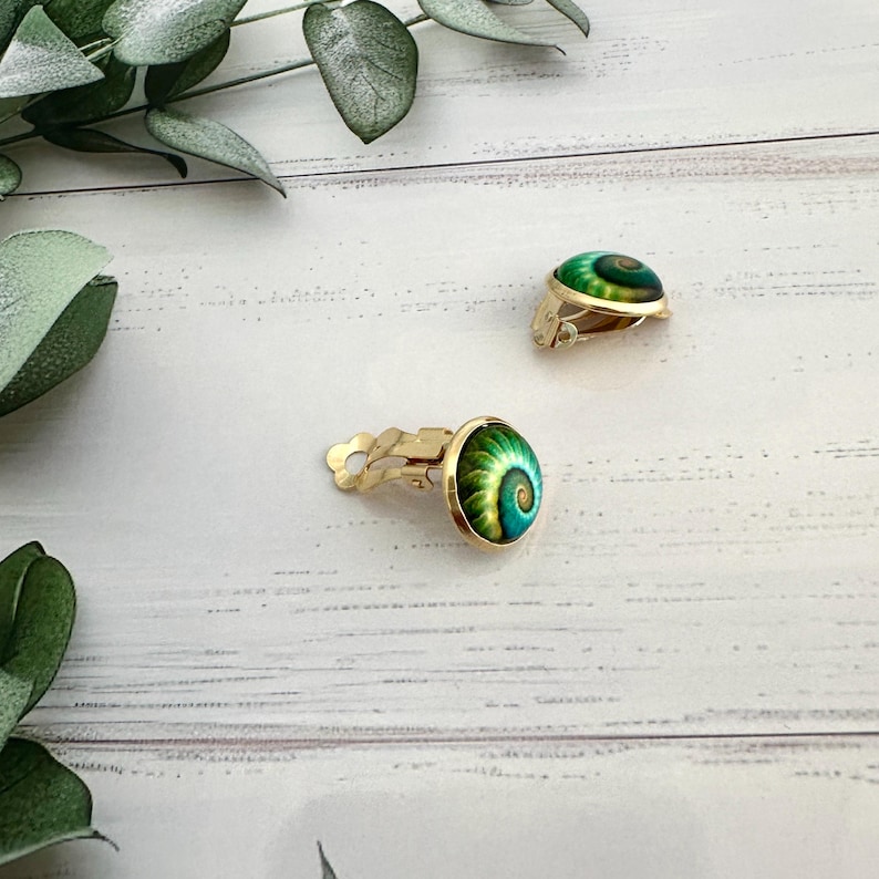 May include: A pair of gold-tone clip-on earrings featuring a circular cabochon with a vibrant green and teal spiral shell pattern. The earrings have a polished gold metal backing and a secure clip mechanism for attachment to the earlobe.