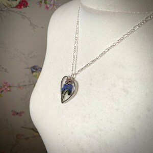 May include: A silver chain necklace with a heart-shaped pendant. The pendant is filled with clear resin and contains a small bouquet of dried blue flowers.