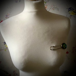 May include: A silver safety pin with a green and black eye charm attached to it. The pin is on a white mannequin torso.