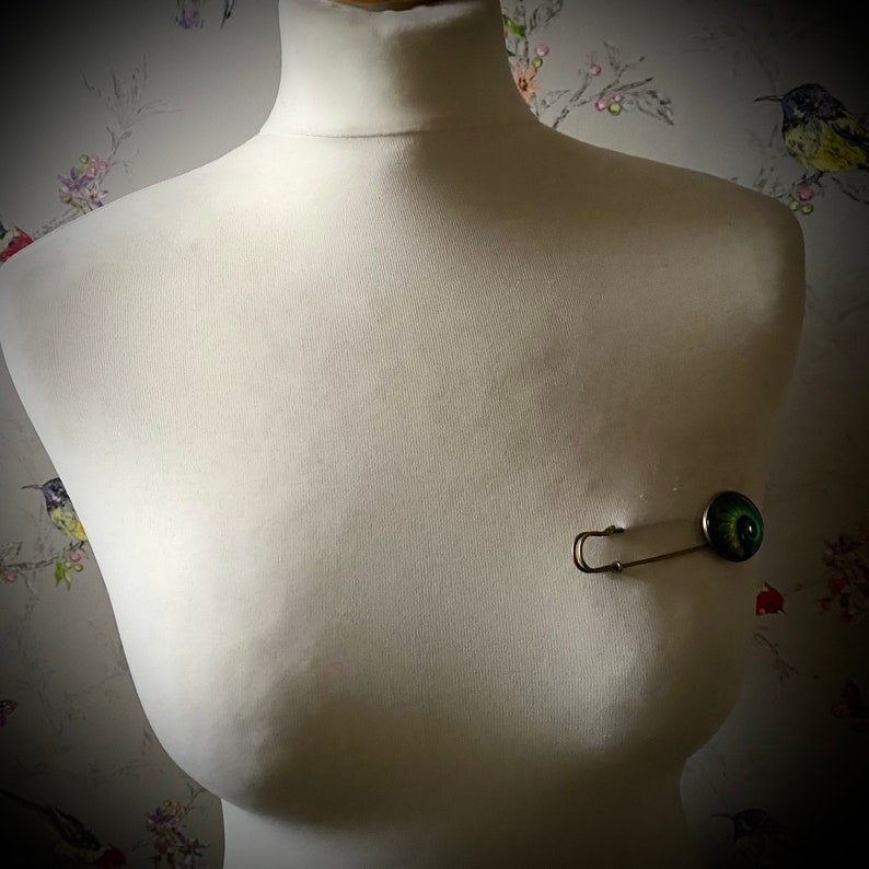 May include: A silver safety pin with a green and black eye charm attached to it. The pin is on a white mannequin torso.