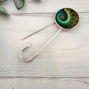 May include: A silver safety pin with a round, green and gold patterned cabochon. The cabochon features a swirling, fractal design.