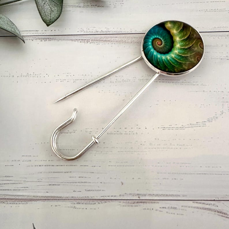 May include: A silver safety pin with a round, green and gold patterned cabochon. The cabochon features a swirling, fractal design.