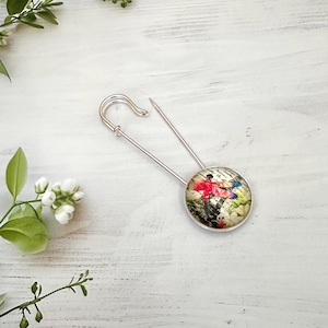 May include: A silver-toned metal safety pin featuring a circular cabochon charm. The charm displays a colourful graphic of a red dragonfly set against a background of vintage-style script text and botanical illustrations in muted tones.