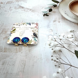 May include: A pair of round clip-on earrings with a gold-tone metal setting and a vibrant blue and purple swirling abstract pattern. The earrings are attached to a white display card featuring the text Marie Appleyard Designs and botanical art.