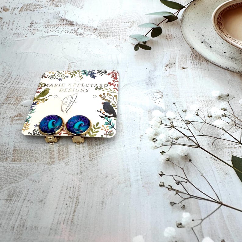 May include: A pair of round clip-on earrings with a gold-tone metal setting and a vibrant blue and purple swirling abstract pattern. The earrings are attached to a white display card featuring the text Marie Appleyard Designs and botanical art.