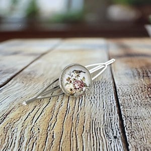 May include: Silver hair clip with a round decorative element. The element features a vintage floral and bird design. The barrette-style clip is designed for securing hair. The image shows the clip on a wooden surface.