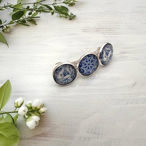 May include: Three silver-toned rings with round, blue and white ceramic inserts. The inserts feature a floral pattern. The rings are arranged on a white wooden surface, with greenery in the background.