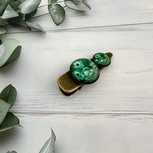 May include: A gold metal hair clip with two round green glass cabochons featuring a peacock feather design.