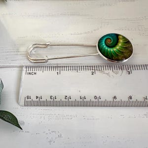 May include: A silver safety pin with a round green, blue, and brown patterned design. The pin is 2 inches long.