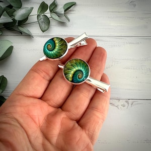 May include: Two silver hair clips with round, green and teal spiral designs. The clips are held in a hand, with a light wood background and green foliage in the upper left corner. The spiral design is reminiscent of a nautilus shell.
