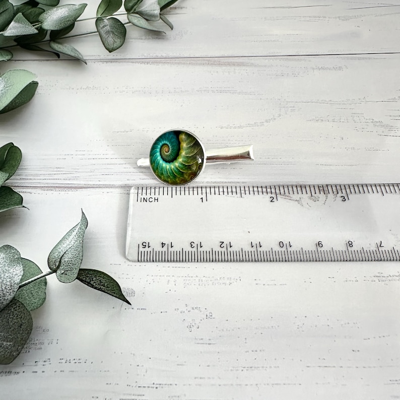 May include: A silver-toned tie clip featuring a circular design with a spiral pattern in shades of green and blue. The tie clip is approximately 1 inch in length and is positioned next to a ruler.