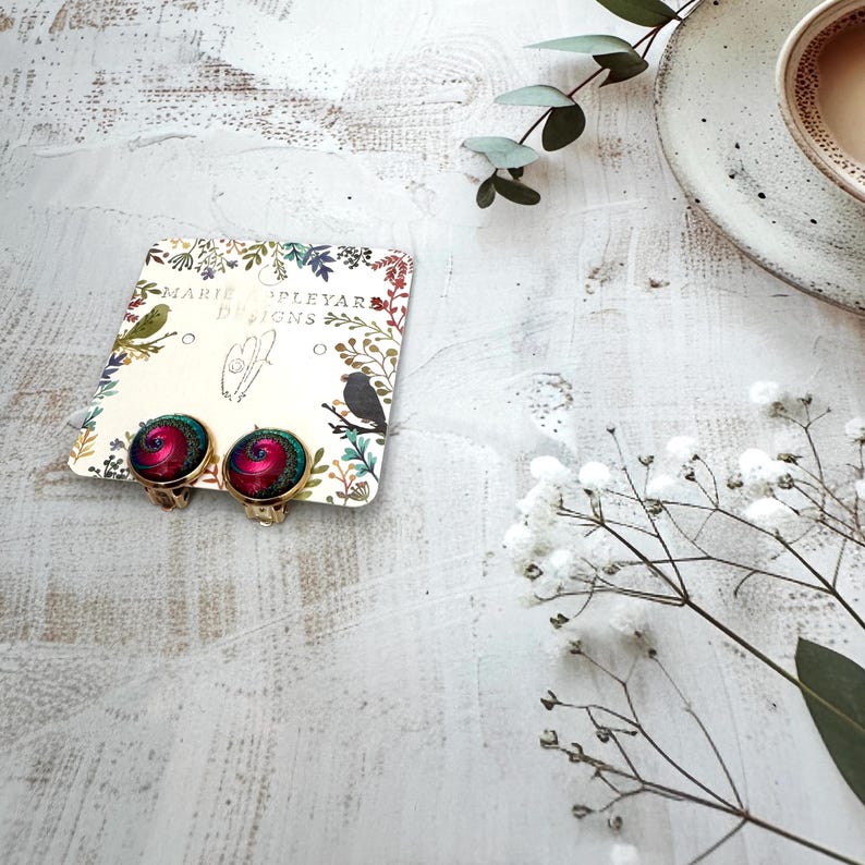 May include: A pair of round clip-on earrings featuring a swirling magenta and teal pattern set in gold-toned metal. The earrings are attached to a cream-colored display card printed with the text Marie Appleyard Designs and botanical illustrations.