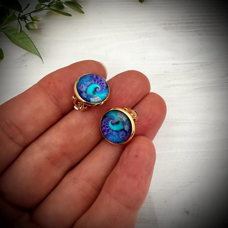 May include: A pair of round gold-tone clip-on earrings featuring a vibrant, swirling fractal pattern in shades of deep purple, blue, and bright turquoise. The circular cabochon centers are set within polished gold-colored metal frames.