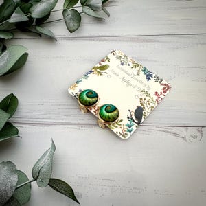 May include: A pair of round clip-on earrings featuring a spiral pattern in shades of green and gold. The earrings are attached to a square display card printed with the text Individual Jewellery by Marie Appleyard Designs and floral motifs.