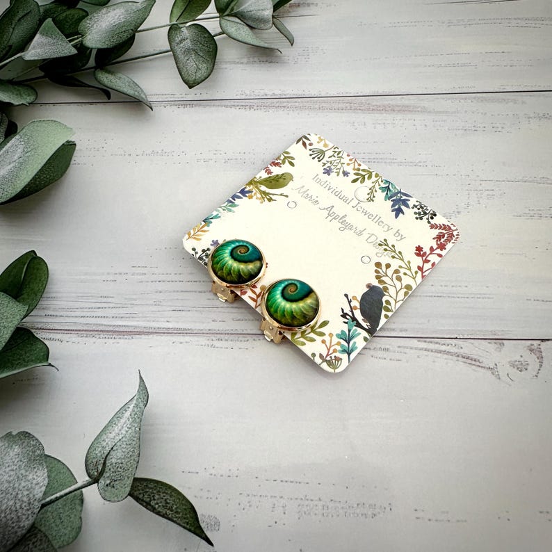 May include: A pair of round clip-on earrings featuring a spiral pattern in shades of green and gold. The earrings are attached to a square display card printed with the text Individual Jewellery by Marie Appleyard Designs and floral motifs.