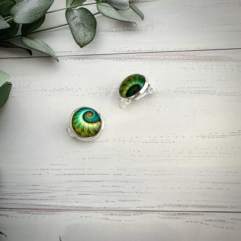 May include: A pair of silver-toned clip-on earrings featuring a circular cabochon with a vibrant green and gold spiral pattern. The earrings have a polished metallic setting and a rounded, dome-shaped surface that displays a detailed nautilus-like design.