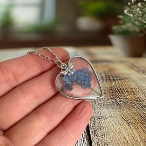 May include: A silver heart-shaped pendant necklace with a delicate chain. The pendant contains a pressed blue forget-me-not flower with a silver bee charm.