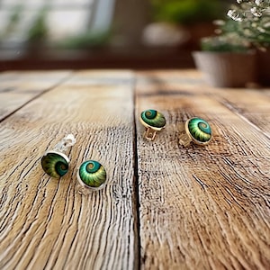 May include: Two pairs of round stud earrings featuring a spiral shell pattern in shades of teal, green, and gold. The earrings have metallic settings and clear plastic backings, displaying a detailed, swirling design on the circular surface.