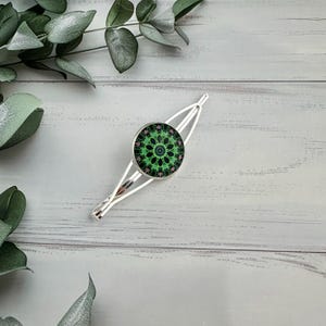 May include: Silver hair clip featuring a circular design with green and pink floral patterns. The clip is set against a light wooden background with green foliage. The clip's design is symmetrical.