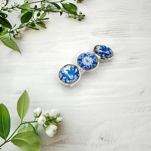 May include: A silver-toned hair clip featuring three circular cabochons with a blue and white pattern. The two outer cabochons display a blue bird motif, while the center cabochon features a blue and white floral or geometric design.