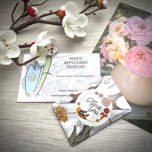 May include: A business card with the text "MARIE APPLEYARD DESIGNS" and a wrapped package with a "Thank You" sticker. The package is wrapped in patterned paper. Decorative flowers and a floral print are in the background.