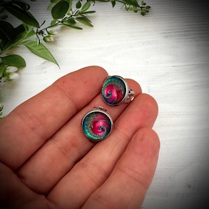 May include: A pair of round stud earrings featuring a metallic silver setting and a vibrant spiral pattern in shades of magenta, pink, and teal. The center of each earring displays a swirling design with a textured, glittery border in dark green.