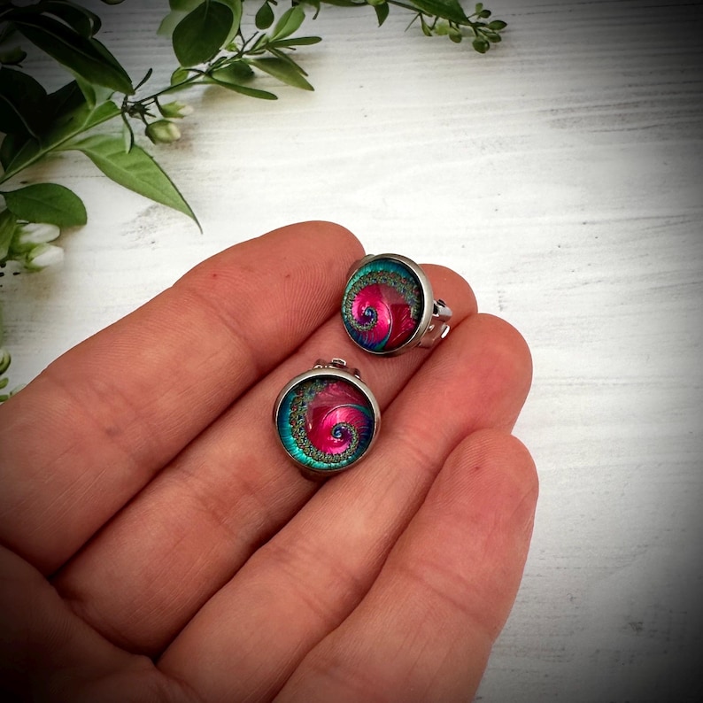 May include: A pair of round stud earrings featuring a metallic silver setting and a vibrant spiral pattern in shades of magenta, pink, and teal. The center of each earring displays a swirling design with a textured, glittery border in dark green.