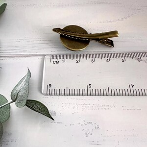 May include: A bronze-colored hair clip with a circular design. The clip is approximately 1 inch long. The clip is next to a ruler with centimeter markings. The clip is on a white surface.