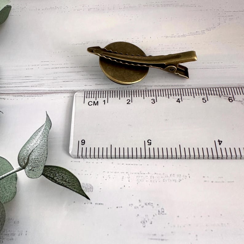 May include: A bronze-colored hair clip with a circular design. The clip is approximately 1 inch long. The clip is next to a ruler with centimeter markings. The clip is on a white surface.