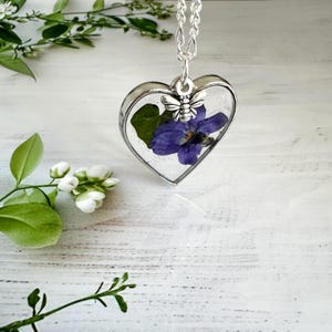 May include: A silver heart-shaped pendant necklace with a pressed purple flower and a silver bee charm. The pendant is filled with clear resin and hangs from a silver chain.