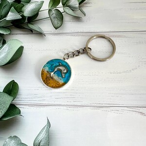 May include: A silver keychain with a round resin charm featuring a dolphin jumping out of blue water with sand at the bottom.