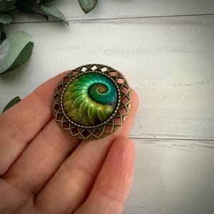 May include: A bronze-coloured pin with a green and blue spiral design inside a geometric frame.
