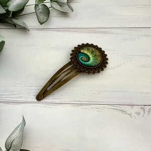 May include: A bronze hair clip with a decorative circular design. The design features a spiral pattern in green and blue, set within a floral-like border. The clip is set against a white wooden surface, evoking a natural theme. The clip is approximately 8 cm long.