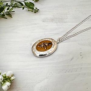 May include: Silver oval locket necklace featuring a bee design. The locket has a floral border and a silver chain. The bee is gold and brown, set against a yellow background. A delicate piece of jewellery.