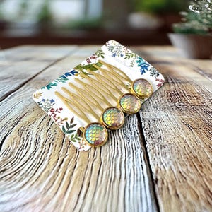 May include: Gold-toned hair combs with iridescent, scale-like circular accents. The combs are arranged on a floral patterned card, creating a whimsical and decorative hair accessory. The combs are designed to be inserted into the hair.