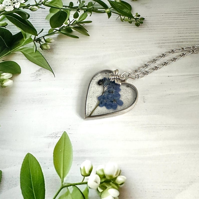 May include: A silver heart-shaped pendant necklace with a delicate silver chain. The pendant contains a pressed forget-me-not flower in clear resin.