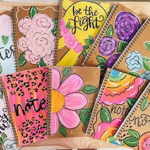 May include: A collection of spiral-bound notebooks with hand-painted floral designs and the word "notes" or "note" in various fonts. The notebooks feature a variety of colors, including pink, yellow, and blue, with black spiral binding.