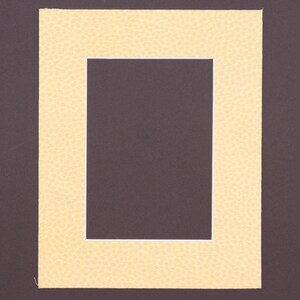 May include: A rectangular picture frame mat with a textured, pale yellow surface. The mat features a central rectangular cutout with a crisp white inner border, framing a dark brown rectangular space in the center of the piece.