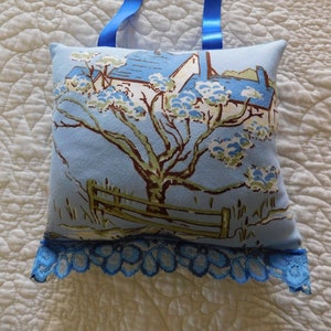 May include: A blue and white pillow with a floral pattern and a house in the background. The pillow has a blue lace trim and a blue ribbon loop for hanging.