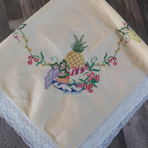 May include: A white tablecloth with a delicate lace trim and an embroidered design of a bowl of fruit, including a pineapple, grapes, and cherries.